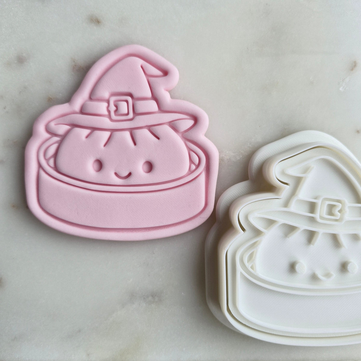 A pink and white cookie cutter shaped like a cute smiling character in a witch hat, displayed on a light marble surface. Halloween Witch Dumpling Cookie Cutter and Stamp