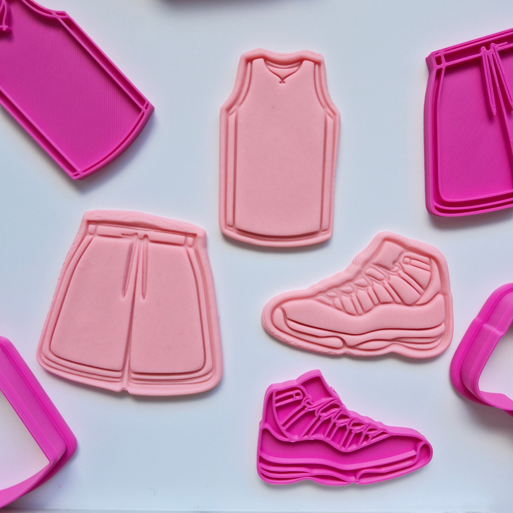 there are 3 pink fondant shapes and cut and stamped by the pink  cookie cutters and stamps making a set of 3 an activewear outfit, includes of jersey single, shorts and sneakers on white background. Activewear Outfit Set Cookie Cutter and Stamp