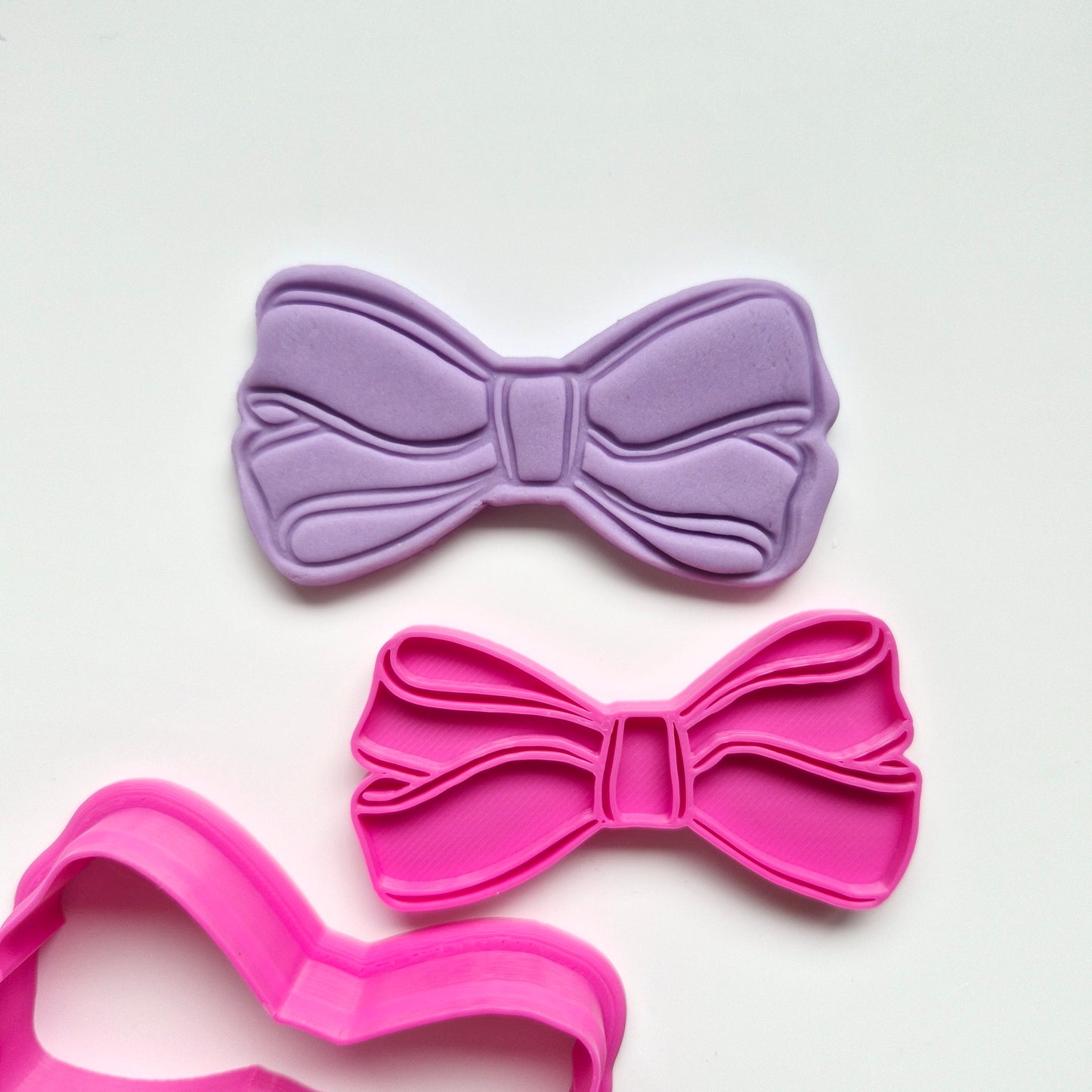 Two bow-shaped cookie cutter stamp and fondant, one purple and one pink, on a white background.
