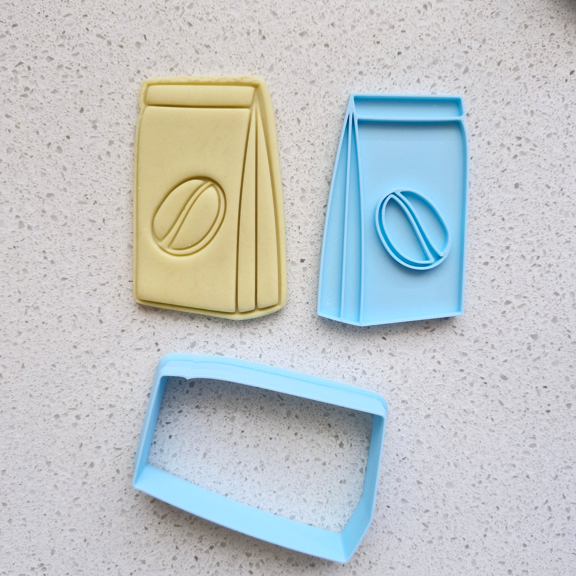yellow fondant, with blue cookie cutter and stamp, in the shape and imprint of a coffee paper bag with espresso bean in the middle, zoomed out, on white speckled stone