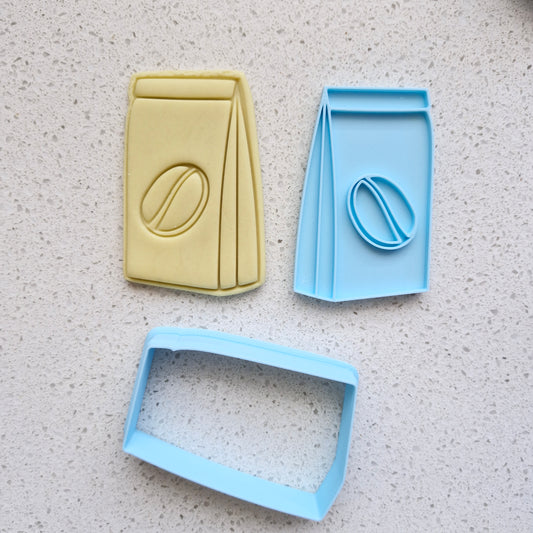 yellow fondant, with blue cookie cutter and stamp, in the shape and imprint of a coffee paper bag with espresso bean in the middle, zoomed out, on white speckled stone