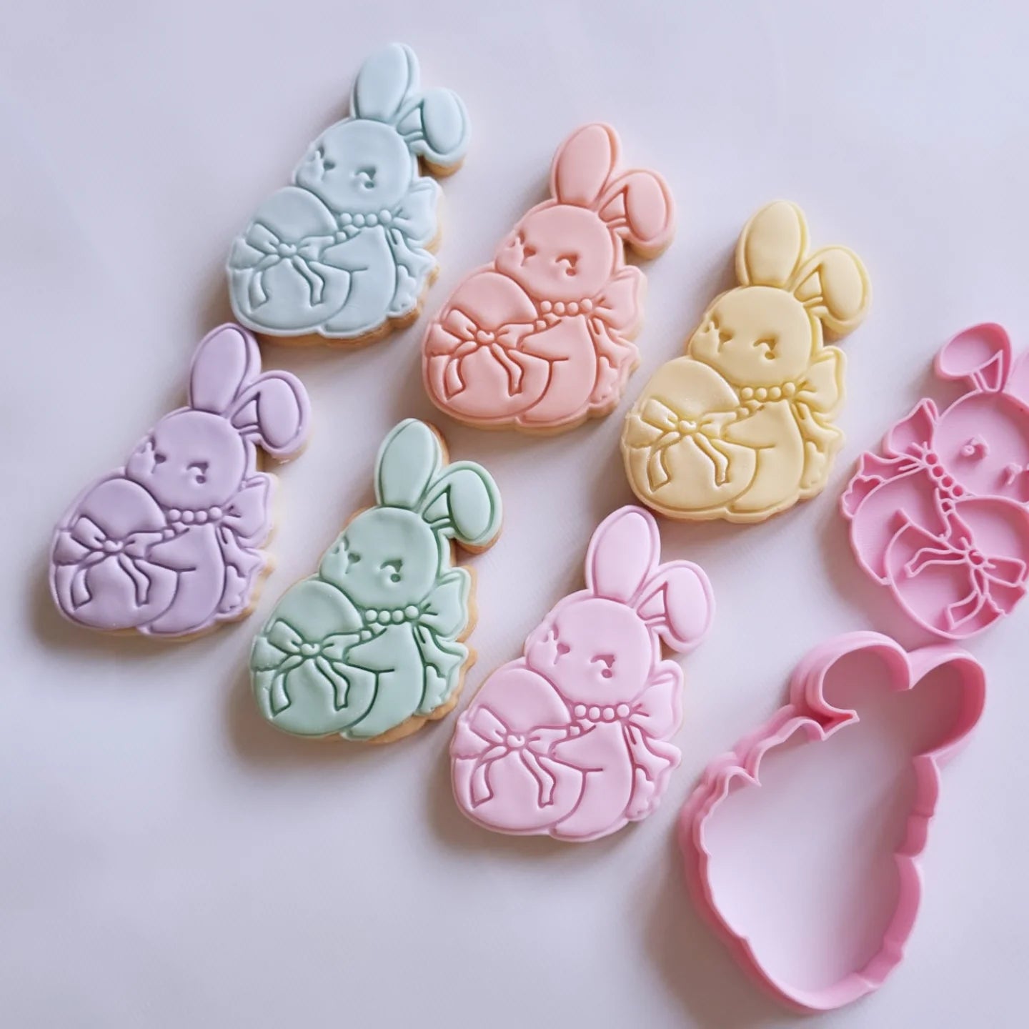 6 sugar cookies pastel colors, the cookies alongside the cookie cutter and stamp are in the design of a coquette bunny holding egg wrapped in a bow Coquette Bunny Cookie Cutter and Stamp
