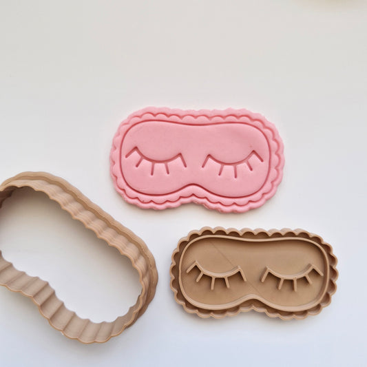 pink fondant, with cookie cutter and stamp, in the shape and imprint of eye mask with closed eyes and eye lashes, on white background zoomed out