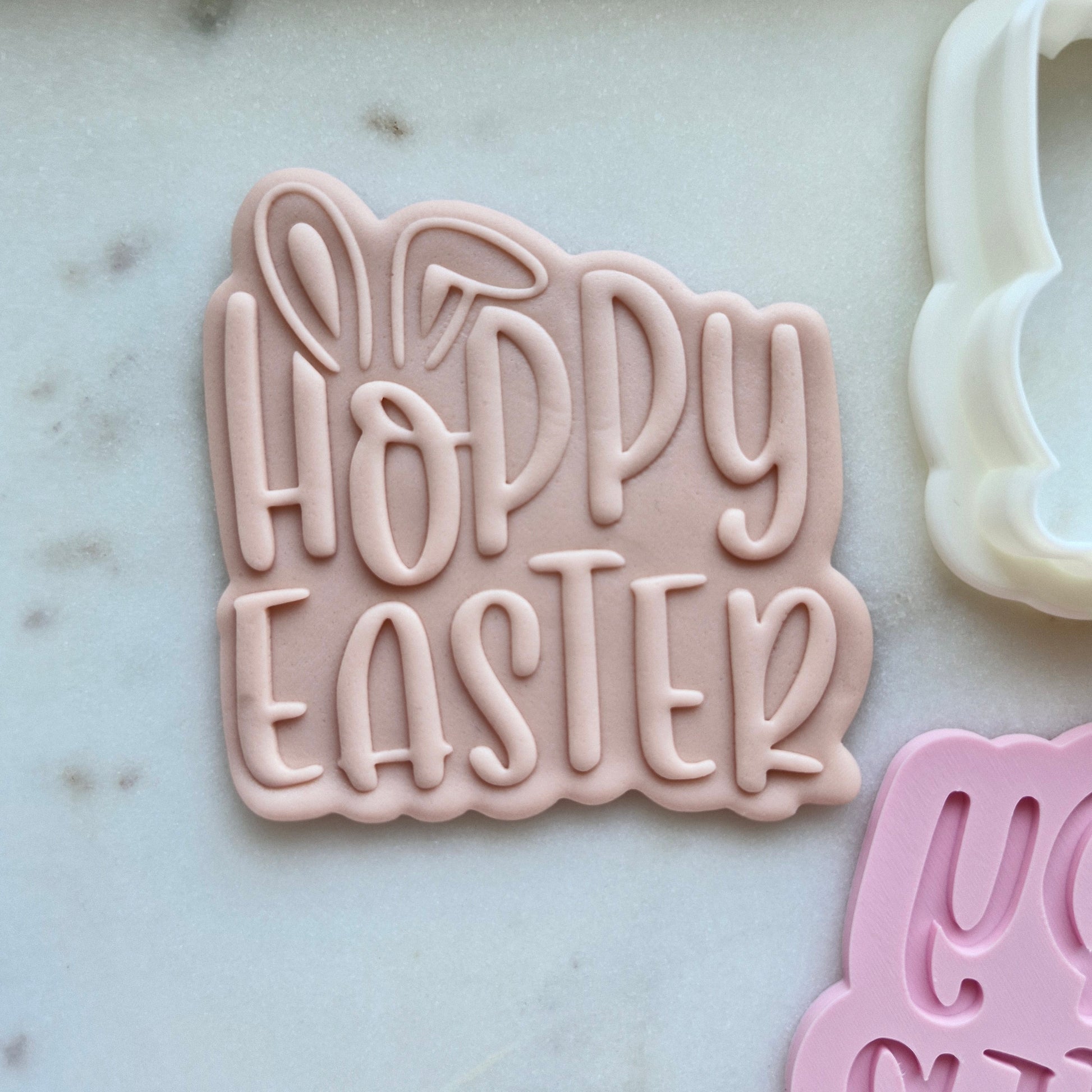 Pink 'Happy Easter' cookie cutter on a marble surface with other cookie cutters in the background.
