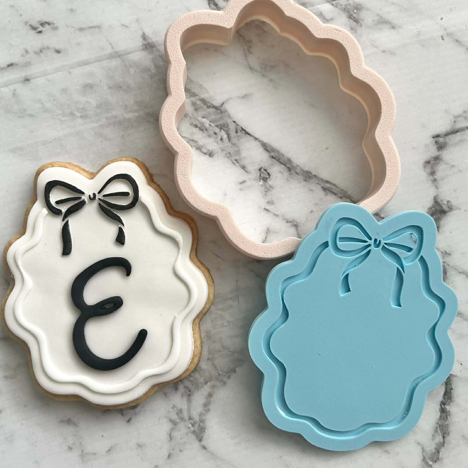 fondant cookie with a cookie cutter and stamp of an oval frame with a bow on the top, the cookie has a custom letter placed on it