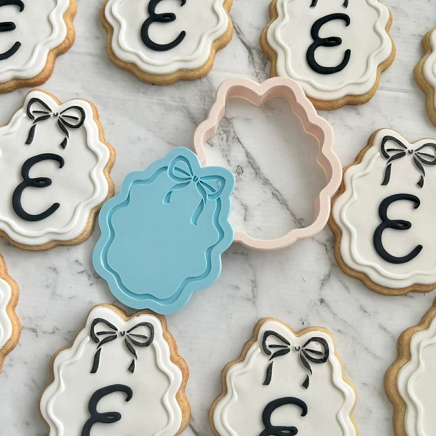 fondant cookie with a cookie cutter and stamp of an oval frame with a bow on the top, the cookie has a custom letter placed on it, with multiple cookie cookies around it