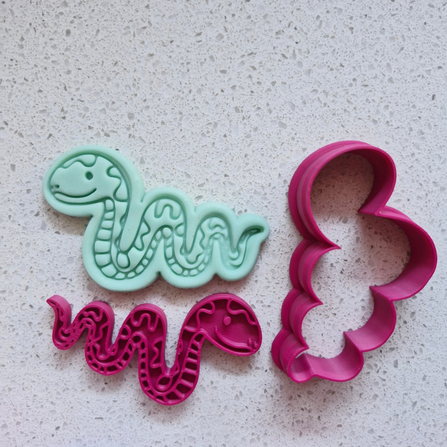 Two snake-shaped cookie cutters, one green fondant and one pink, on a textured surface of speckled stone Slithering