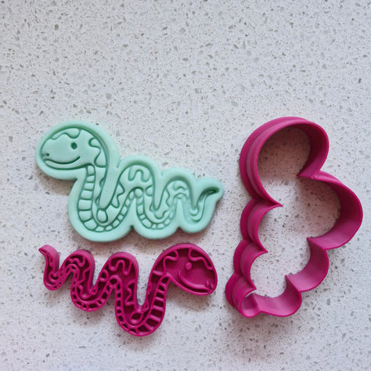 Two snake-shaped cookie cutters, one green fondant and one pink, on a textured surface of speckled stone Slithering